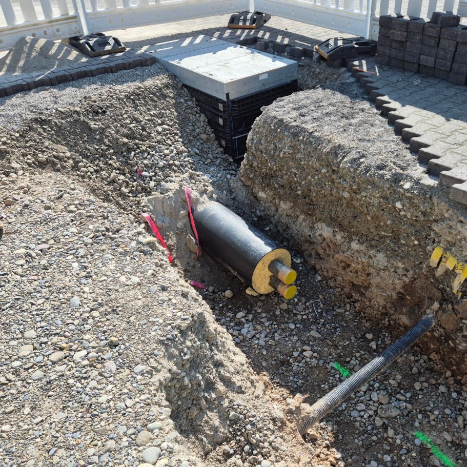 Offfener Tiefbaugraben mit laufenden Rohrleitungsarbeiten für die Gewerke Nahwärme, Wasser, Breitband und Entwässerung.