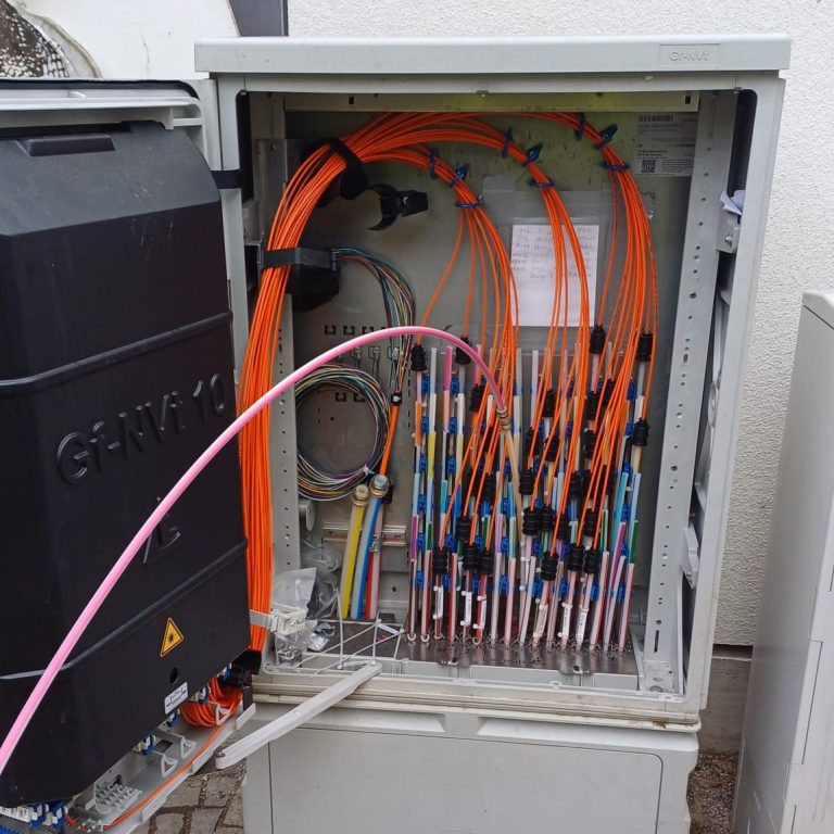 Ein geöffneter Glasfaser Netzverteiler mit orangenen Kabeln und Anschlussleitungen in einer städtischen Umgebung.